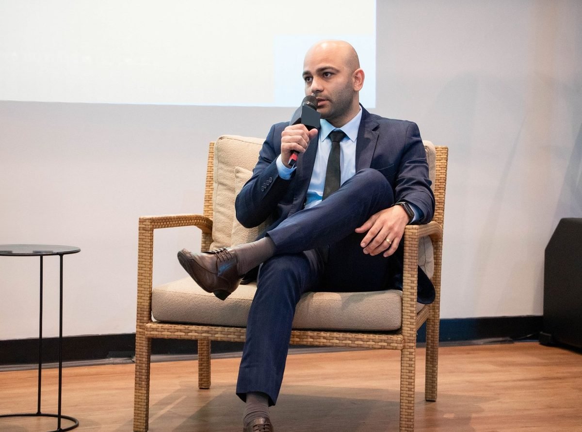 Caio Nascimento numa poltrona, falando ao microfone, em luz natural
